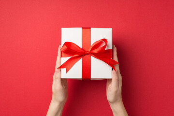 First person top view photo of young woman's hands holding big white gift box with red ribbon bow on isolated red background