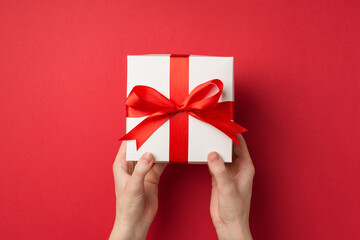First person top view photo of woman's hands giving big white giftbox with red silk ribbon bow on isolated red background