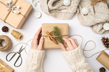 Fototapeta na wymiar First person top view photo of hands in sweater decorating craft paper giftbox with fir twig cinnamon stick dried lemon slice scarf christmas tree balls and handicraft tools isolated white background
