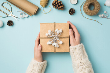 Fototapeta na wymiar First person top view photo of female hands in knitted pullover holding giftbox with snow twig balls scissors spools of twine roll of craft paper candle and cones on isolated pastel blue background