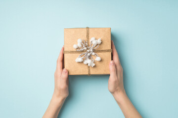 First person top view photo of female hands holding craft paper giftbox decorated with snow twig and twine on isolated pastel blue background