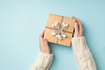First person top view photo of female hands in white sweater demonstrating kraft paper gift box decorated with snow twig and twine on isolated pastel blue background