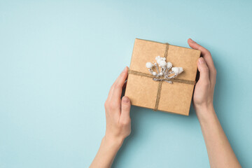 First person top view photo of girl's hands holding kraft paper gift box decorated with snow twig and twine on isolated pastel blue background with copyspace