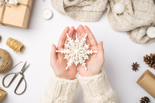 First person top view photo of female hands in sweater holding big decorative snowflake giftbox scarf christmas tree balls cones anise candles scissors spools of twine on isolated white background