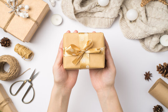 First person top view photo of young woman's hands holding craft paper giftbox with gold ribbon bow over sweater christmas tree balls and handicraft tools on isolated white background