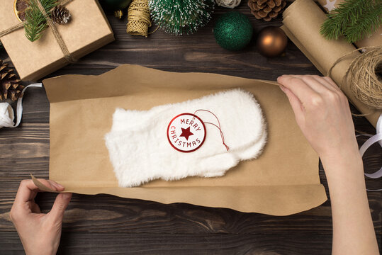 First person top view photo of woman's hands packing white fluffy mittens with tag merry christmas in kraft paper green gold balls pine twigs and handicraft tools on isolated wooden desk background