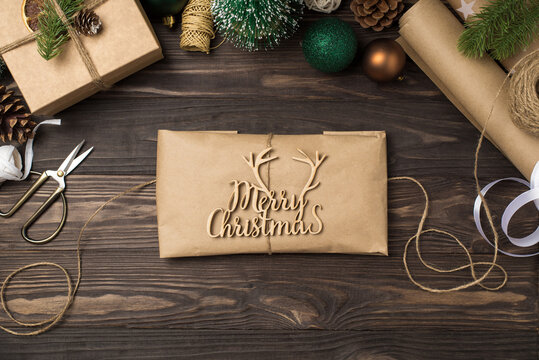 Top view photo of craft paper package with merry christmas wooden text and twine green gold balls pine twigs cones giftbox scissors paper rolls and spools on isolated dark wooden desk background