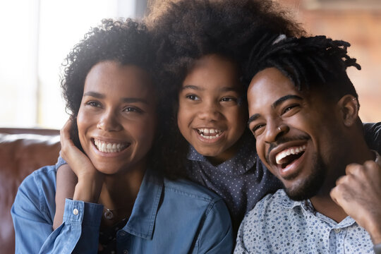 Head shot happy bonding African American two generations family parents resting with little cute child daughter on cozy sofa, looking in distance, watching funny smart TV programs or shows at home.