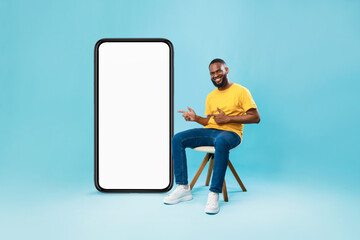 Happy black guy sitting on chair, pointing at giant smartphone with white screen, offering mockup for mobile app design