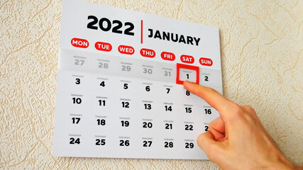 Close-up of the first page of a wall calendar 2022 and a male hand moves a red cursor to the 1st...
