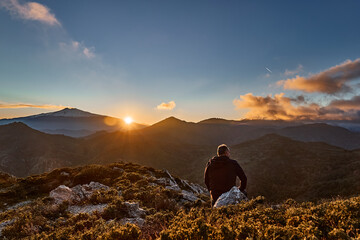 Man sitting on top of the cliff in the mountains at sunset enjoying the beautiful sunset over...