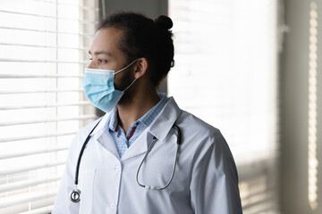 Profile close up pensive African American man doctor in medical mask thinking, looking at window in hospital, confident thoughtful physician practitioner in white uniform planning, visualizing