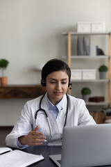 Vertical photo of young Indian woman doctor in headphones talking, using laptop in office, female...