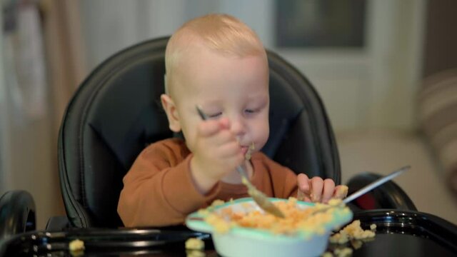 A child with white hair and a brown T-shirt is sitting in a black children's chair and eating yellow porridge