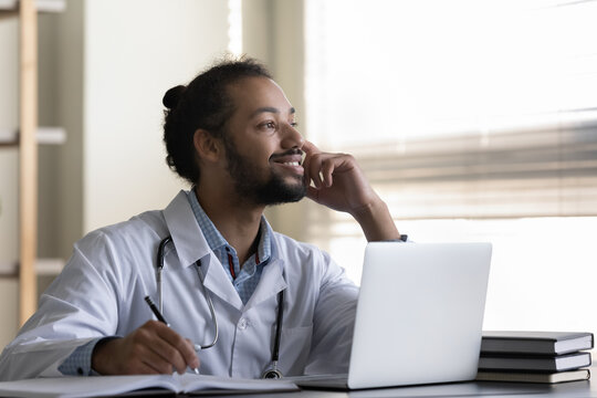 Pensive Smiling Young African American Man Doctor Or Student Intern Thinking, Studying Online In Office, Taking Notes, Using Laptop, Dreamy Physician Visualizing Opportunities Or Future, Planning