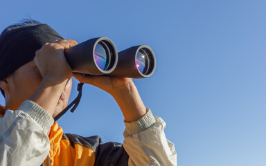 Young male man looking through the binoculars on mountain at beautiful blue sky background.