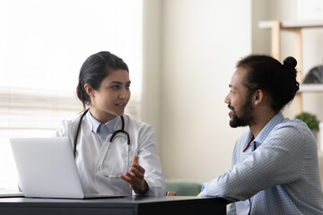 Fototapeta premium Smiling Indian woman doctor consulting African American man patient, therapist practitioner giving recommendation, discussing checkup results or symptoms at meeting, sitting at desk with laptop