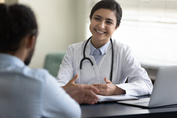 Smiling young Indian woman doctor consulting African American patient at meeting in hospital, friendly female physician practitioner talking, giving recommendation, discussing checkup or symptoms