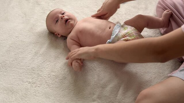 Health skin care, development pediatrics, infant close-up mother hands give naked newborn baby child massage on arms, legs, fingers, reflex on feet exercise, rubbing skin abdomen chest on bed at home