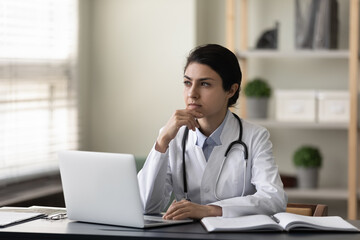 Thoughtful serious Indian female doctor physician touching chin sitting at work desk in office with...