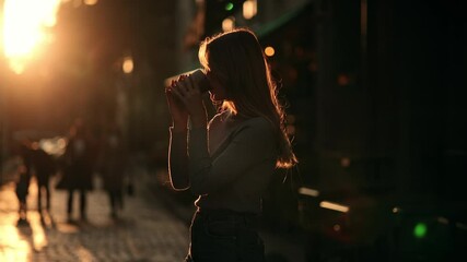 Silhouette of Happy young hipster woman drinking coffee at sunrise. Girl taking a sip of coffee or tea from wooden eco cup on warm summer evening sunset in big city. Natural beauty pretty woman