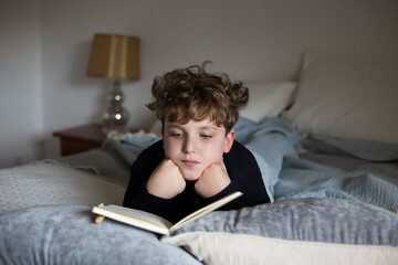 Cute ten-year old boy reading book while stretched out on bed at home