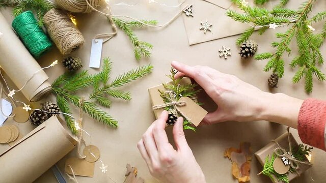 Hands pack a gift for Christmas and new year in eco-friendly materials: kraft paper, live fir branches, cones, twine, tied with a bow. Tags with mock up, natural decor, hand made, flatly. Festive mood