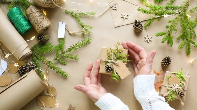 Hands pack a gift for Christmas and new year in eco-friendly materials: kraft paper, live fir branches, cones, twine, tied with a bow. Tags with mock up, natural decor, hand made, flatly. Festive mood