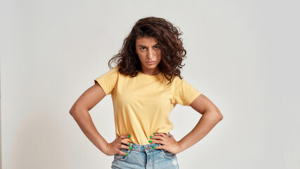 Confident young female model with dark curly hair in casual wear looking at camera while posing isolated over gray background