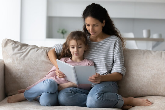 Caring mother with adorable 7s daughter reading book together, sitting on couch at home, loving mom teaching little girl to read, family engaged in educational activity, enjoying fairy tale story - Powered by Adobe
