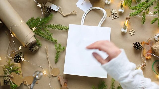 Hands pack a gift for Christmas and new year in eco-friendly materials: kraft paper, live fir branches, cones, twine, tied with a bow. Tags with mock up, natural decor, hand made, flatly. Festive mood