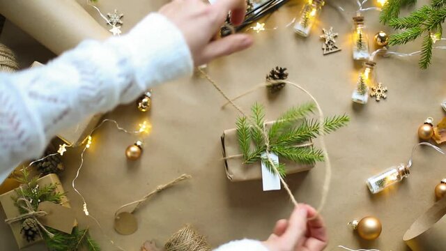Hands pack a gift for Christmas and new year in eco-friendly materials: kraft paper, live fir branches, cones, twine, tied with a bow. Tags with mock up, natural decor, hand made, flatly. Festive mood