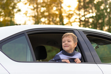 A seven year old cute boy leans out the window of a white car on a warm sunny autumn day against the backdrop of yellow foliage. Selective focus. Portrait
