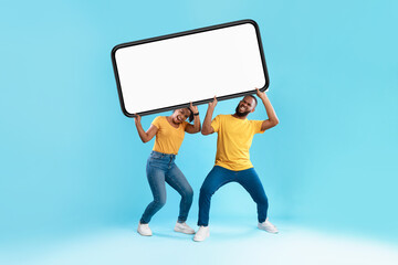 Emotional black couple holding big heavy smartphone with empty screen above their heads on blue...
