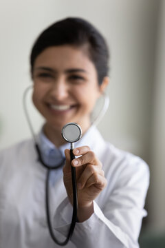 Vertical photo close up smiling Indian female doctor physician in white uniform holding stethoscope, examine checking patient, focus on medical equipment in practitioner hand, healthcare concept