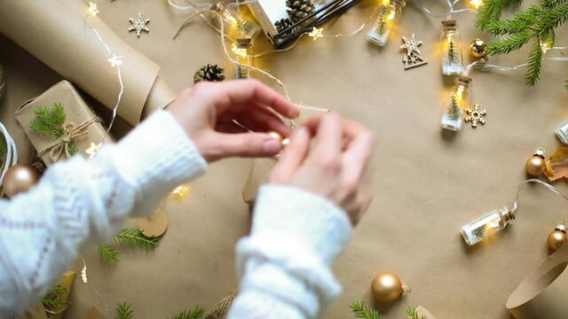 Hands pack a gift for Christmas and new year in eco-friendly materials: kraft paper, live fir branches, cones, twine, tied with a bow. Tags with mock up, natural decor, hand made, flatly. Festive mood