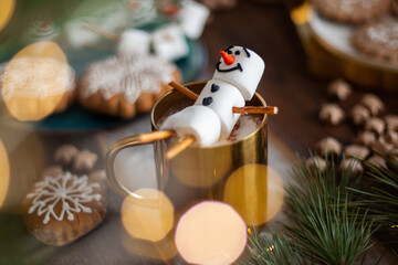 A marshmallow snowman decorated with icing. Gold mug with cocoa and Christmas decor. Gingerbread in...