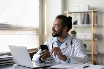 Fototapeta na wymiar Smiling pensive African American man doctor wearing white uniform with stethoscope holding smartphone sitting at desk with laptop, looking in distance, planning and visualizing, distracted from phone