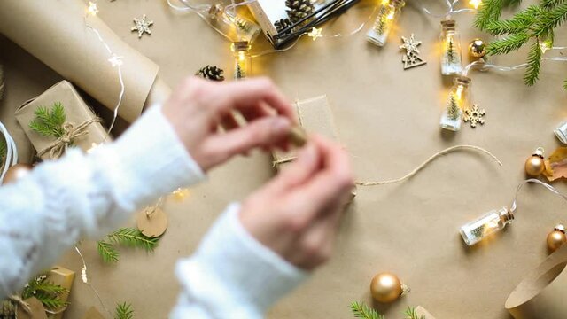 Hands pack a gift for Christmas and new year in eco-friendly materials: kraft paper, live fir branches, cones, twine, tied with a bow. Tags with mock up, natural decor, hand made, flatly. Festive mood