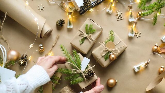 Hands pack a gift for Christmas and new year in eco-friendly materials: kraft paper, live fir branches, cones, twine, tied with a bow. Tags with mock up, natural decor, hand made, flatly. Festive mood