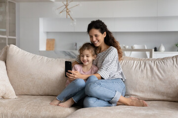 Happy family mother and little daughter having fun with modern smartphone, sitting on couch at home, smiling mom with cute girl kid chatting online by video call, taking selfie, watching cartoons