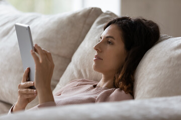 Close up profile of woman using tablet device, relaxing on couch at home, beautiful young female...