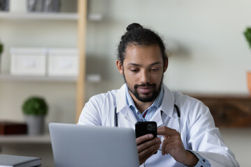 Confident young African American man doctor physician in white uniform using smartphone in office, looking at phone screen, reading message, consulting patient online, browsing healthcare apps