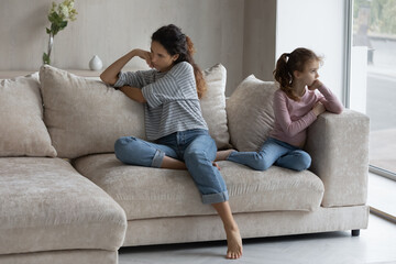 Fototapeta premium Unhappy mother and stubborn little daughter ignoring each other after quarrel, sitting on couch at home back to back, child and parent conflict concept, family misunderstanding and generation gap