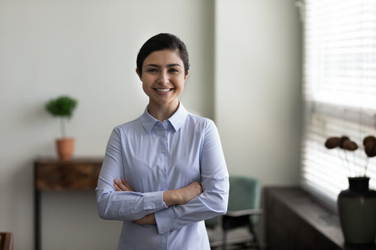 Head shot portrait smiling Indian woman with folded hands standing in modern office room, successful confident businesswoman entrepreneur looking at camera, posing for corporate profile picture