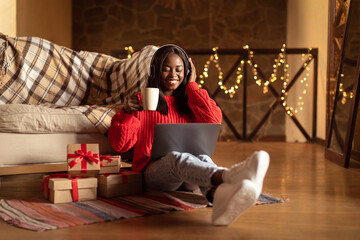 Happy black woman in headphones using laptop, drinking coffee, sitting on floor near Christmas gift boxes at home