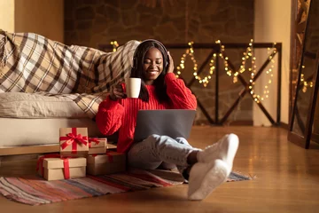 Photo sur Plexiglas Magasin de musique Happy black woman in headphones using laptop, drinking coffee, sitting on floor near Christmas gift boxes at home