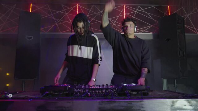 Energetic attractive multiracial male djs in headphones performing live set at stage, mixing music on modern sound mixer and turntables, motivating and cheering up people in nightclub.