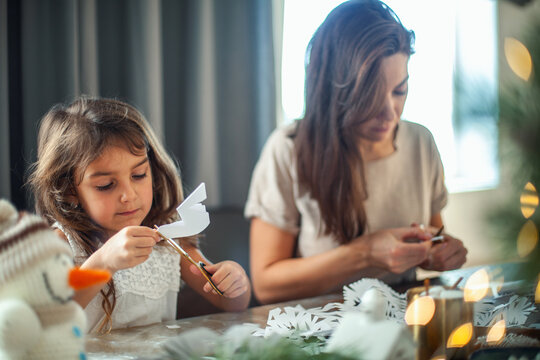 Little cute girl and young beautiful woman cut snowflakes from white paper. Gingerbread and cocoa with marshmallows.