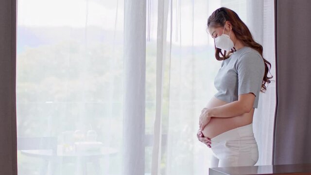 Happy Pregnant Woman wear medical face mask protect virus standing in front of windows stroking big belly with love at cozy home, Pregnancy of young woman enjoy with future life